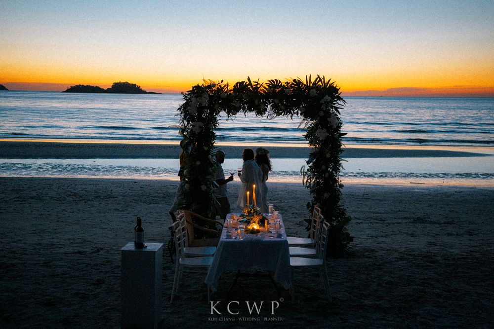Romantic beach dinner setup at sunset with floral arch and candlelit table on the sand overlooking the ocean.