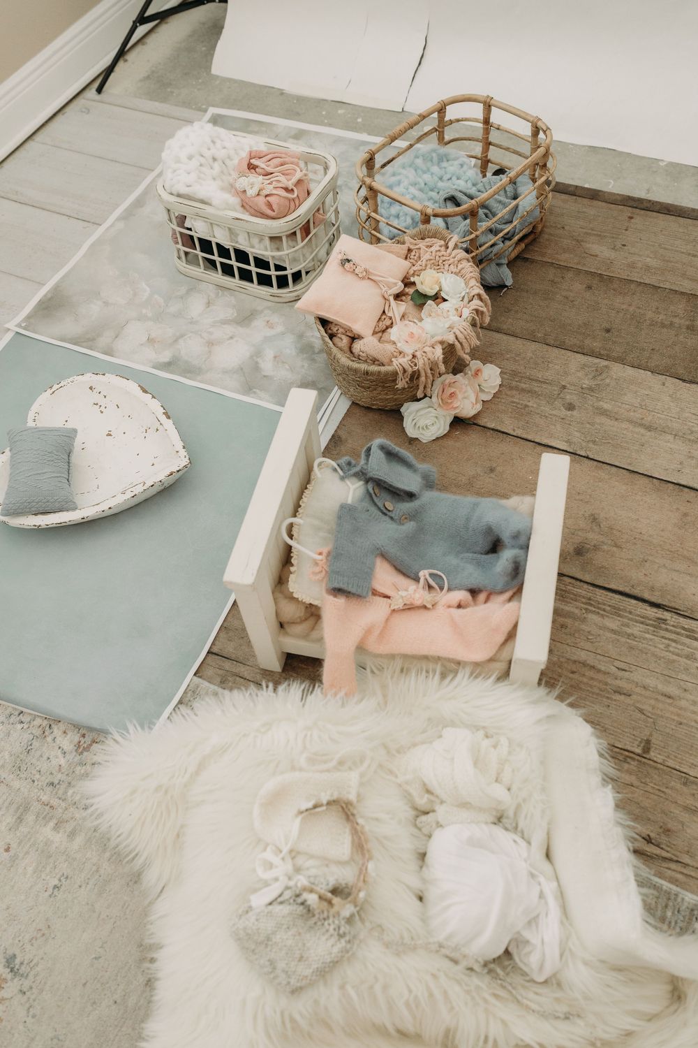 Soft pastel baby items including baskets, blankets, and clothing arranged on a rustic wooden floor.