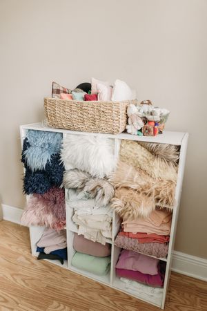 Organized storage unit with plush blankets, throws and pillows arranged neatly in white cubby shelves with woven basket on top.