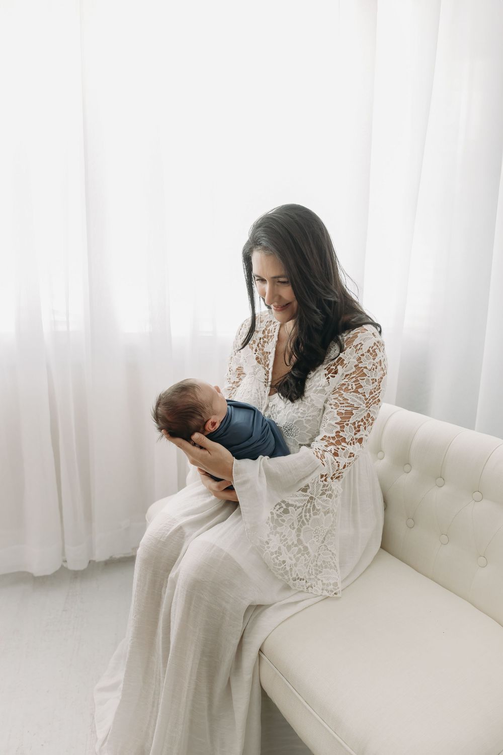 Woman in white lace dress sitting on cream sofa holds small infant in intimate indoor portrait setting.