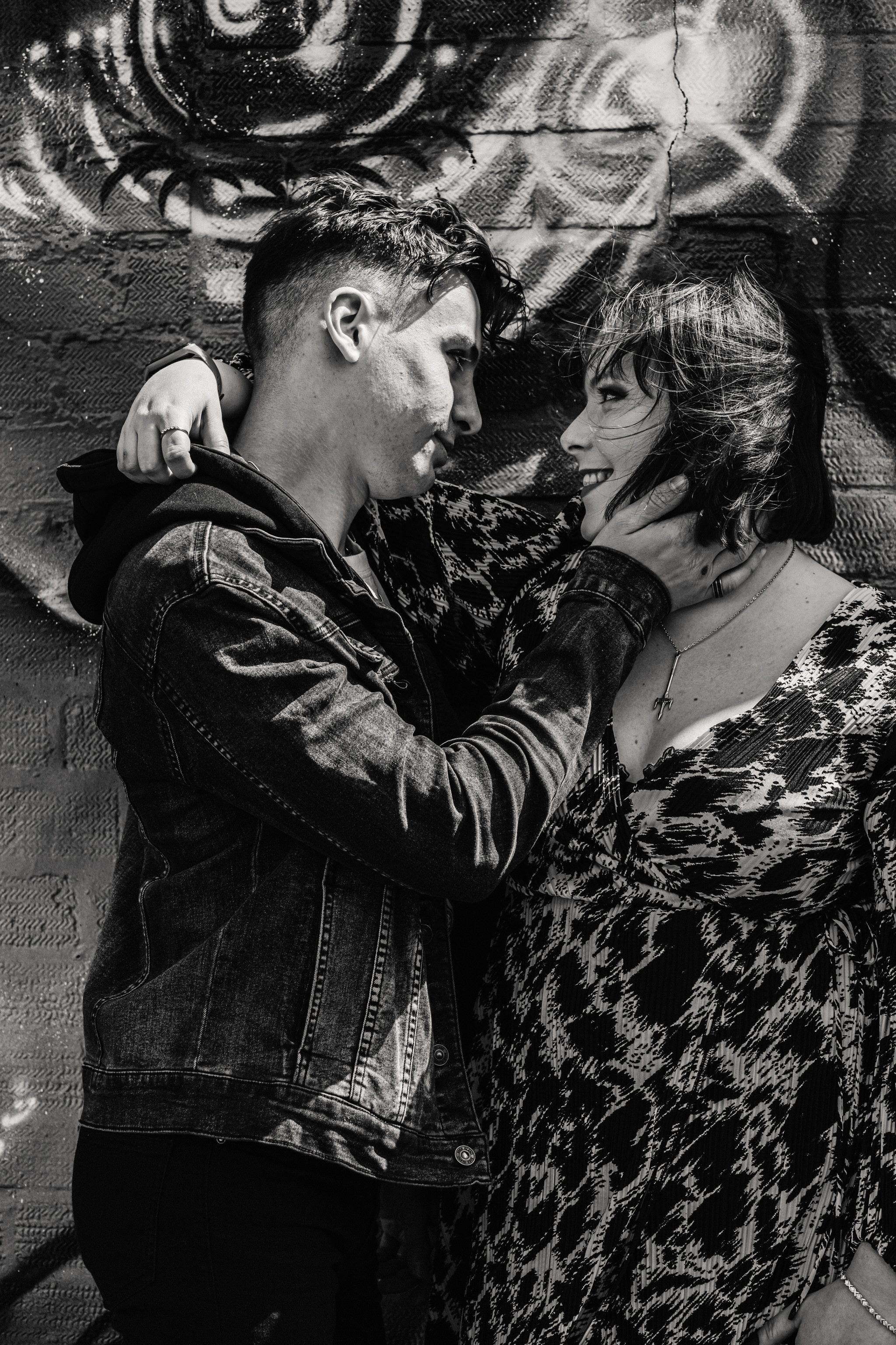 A couple in black and white embraces against a graffiti wall in an urban setting with an artistic black eye mural.