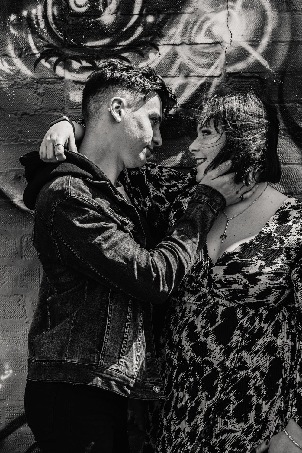 A couple in black and white embraces against a graffiti wall in an urban setting with an artistic black eye mural.