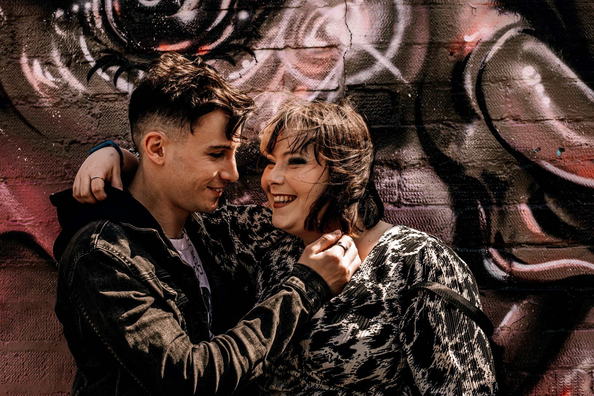 Couple in edgy outfits share tender moment against urban graffiti wall with pink and dark tones.