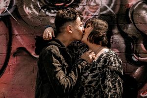 A romantic couple shares a tender kiss against a graffitied red wall background.