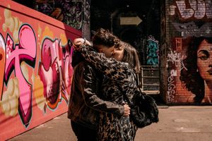 People embrace in front of vibrant graffiti walls with pink street art and an arrow sign visible in an urban setting.