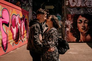 A couple embraces in front of vibrant street art and graffiti on brick walls in an urban setting.