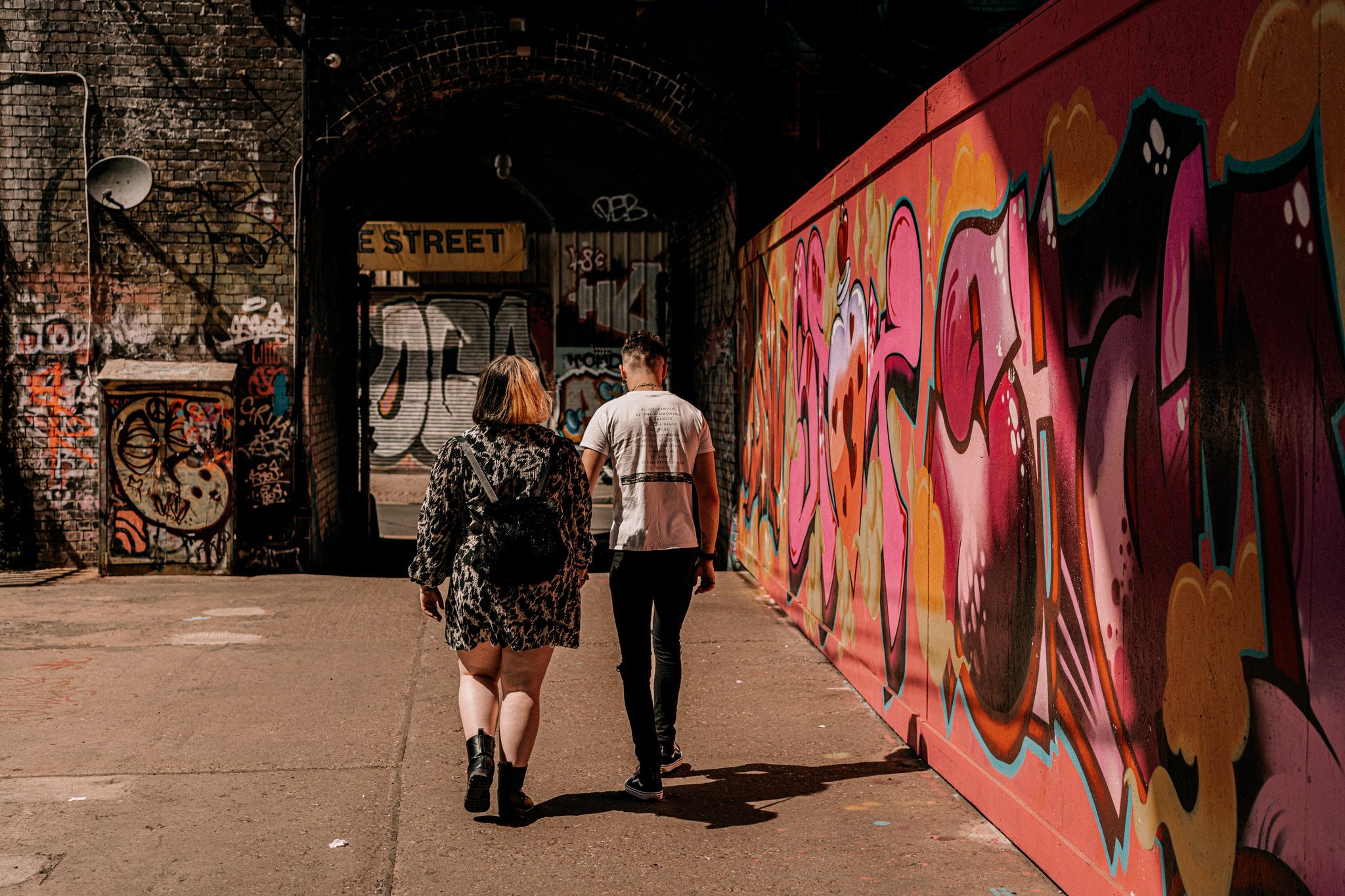 Two people walk past vibrant pink graffiti murals on an urban street wall in warm sunlight.