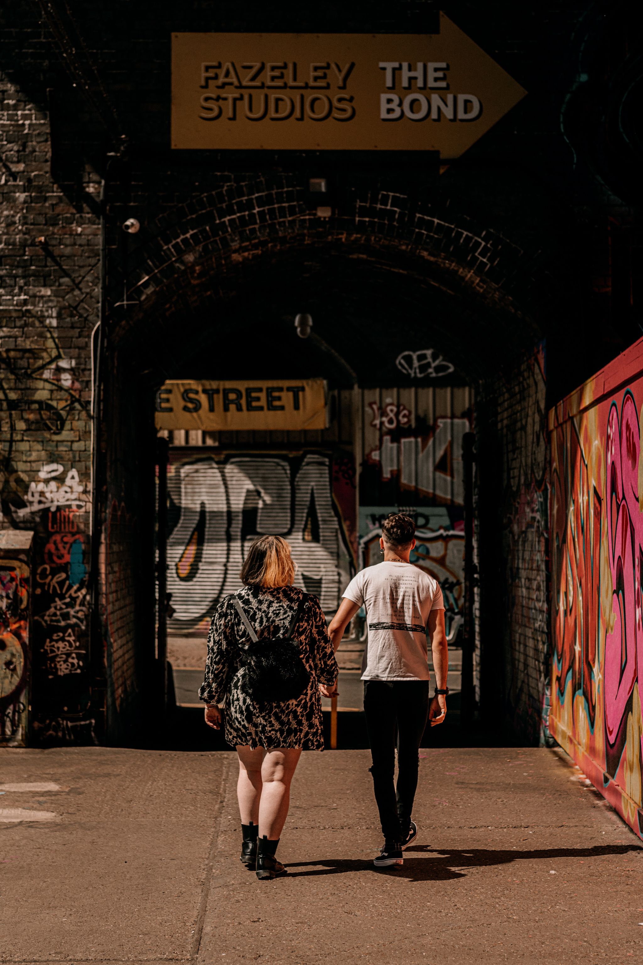 Two people walking under gritty urban archway with graffiti and street art at Fazeley Studios in Birmingham.