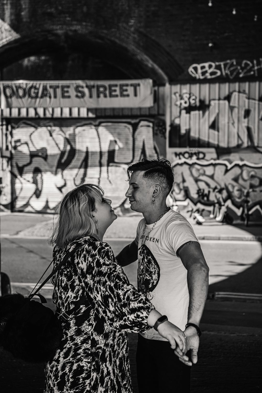 Black and white photo of a romantic couple laughing together under a graffitied bridge on Drygate Street.