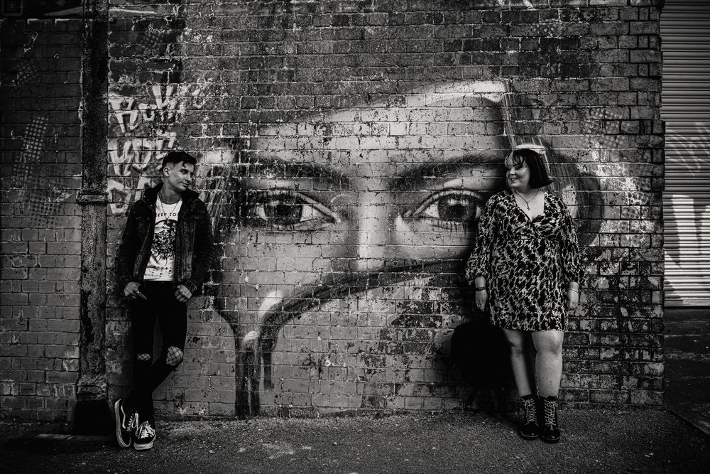 Black and white street art mural of large eyes painted on brick wall with two people posing beside it.