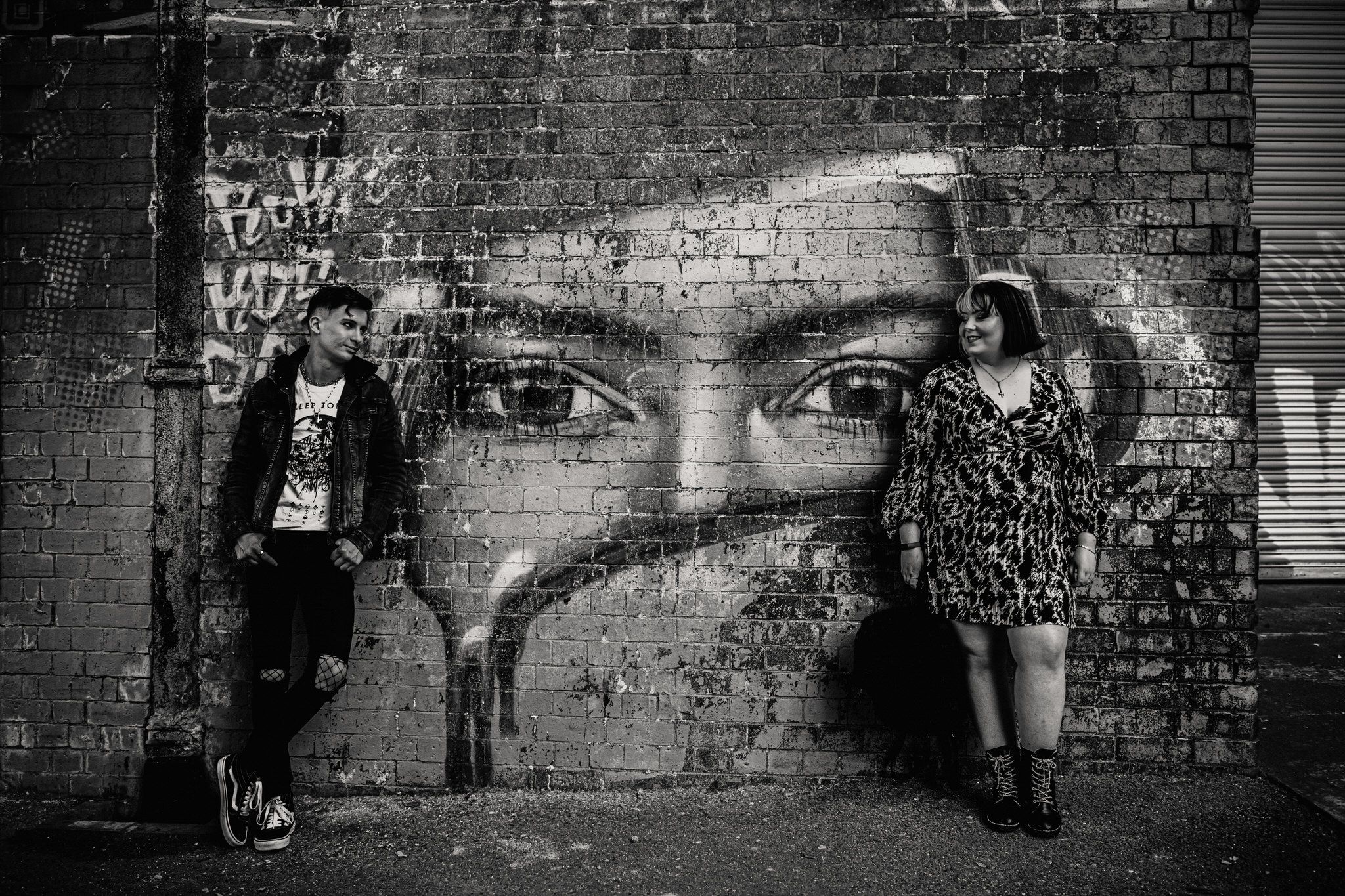 Black and white street art mural of large eyes painted on brick wall with two people posing beside it.