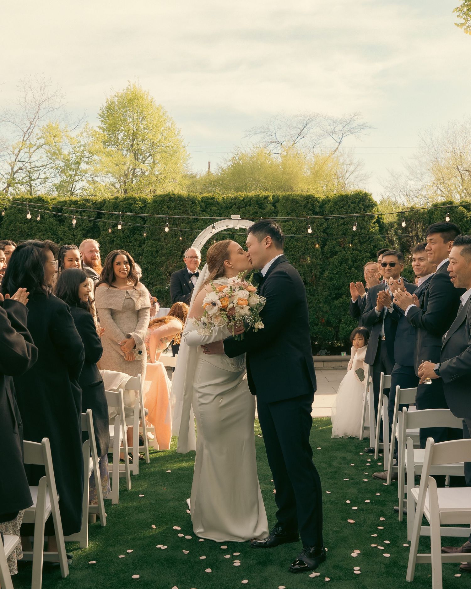 Outdoor wedding ceremony with string lights and white chairs on grass while guests look on during celebration.