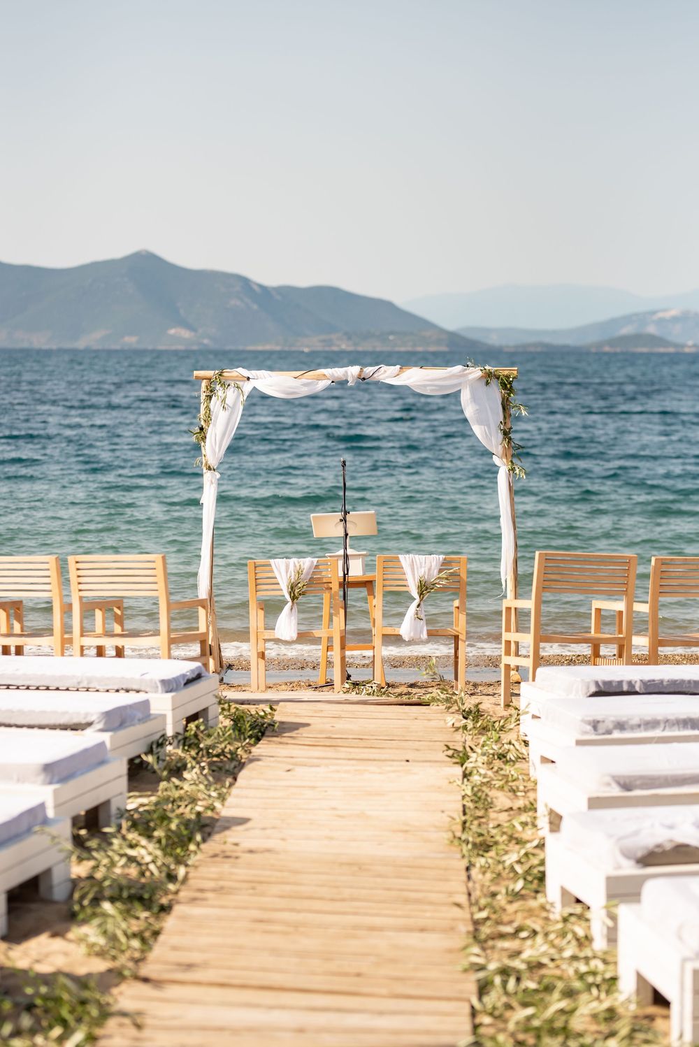 Seaside wedding ceremony setup with white chairs and floral arch overlooking blue waters and mountains.