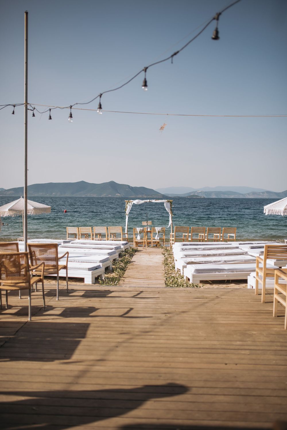 A beachfront wedding ceremony setup with white benches and a floral arch overlooking mountains and ocean under string lights.