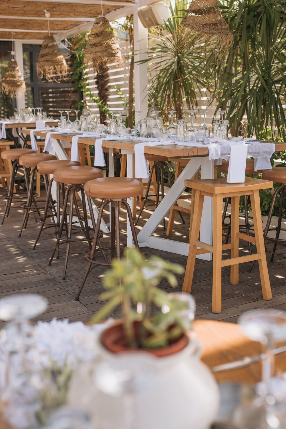 Modern outdoor restaurant seating with wooden barstools and white tables decorated with plants in a sunny patio setting.