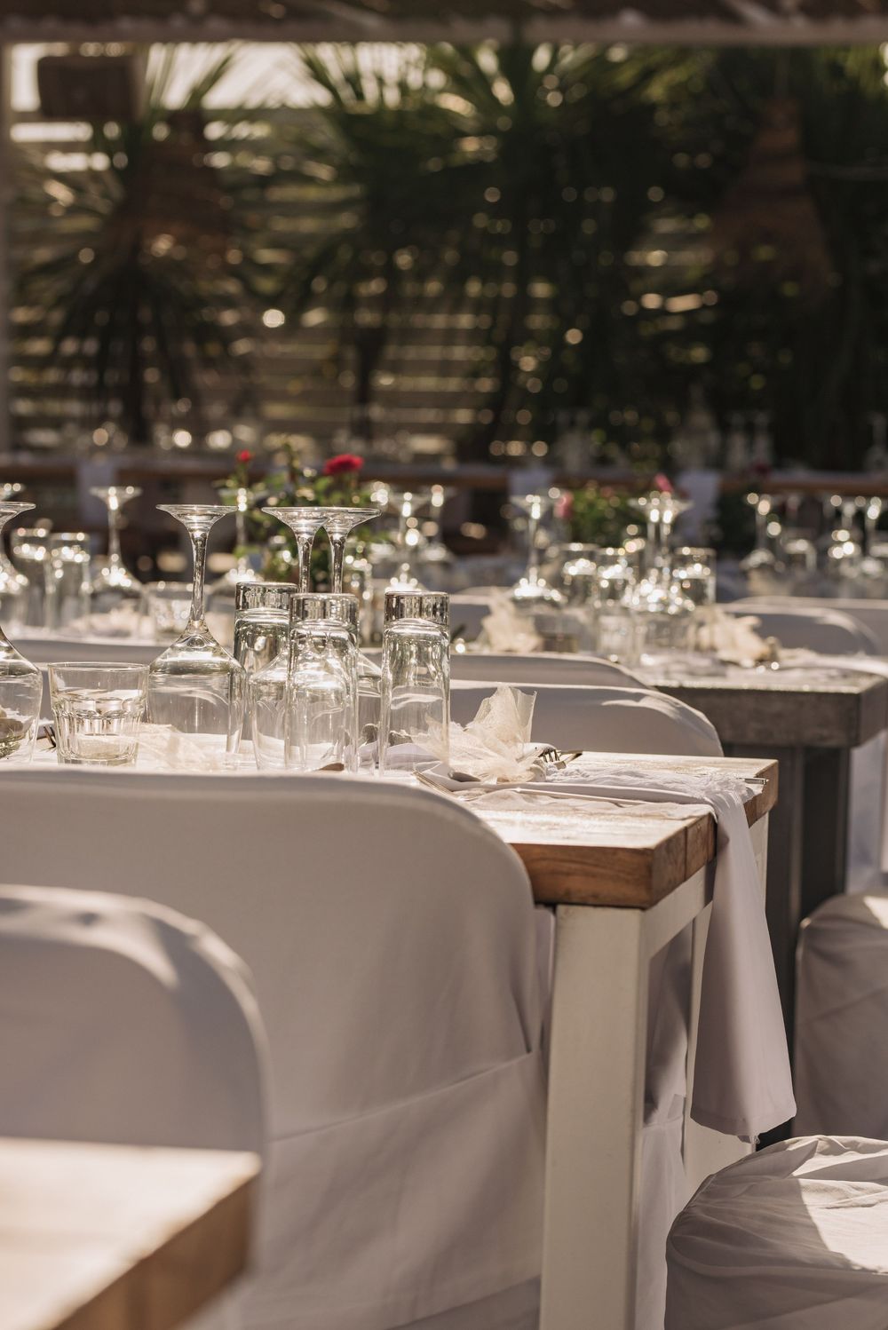 Elegant wedding reception table setting with white linens, glassware and palm fronds visible in background.