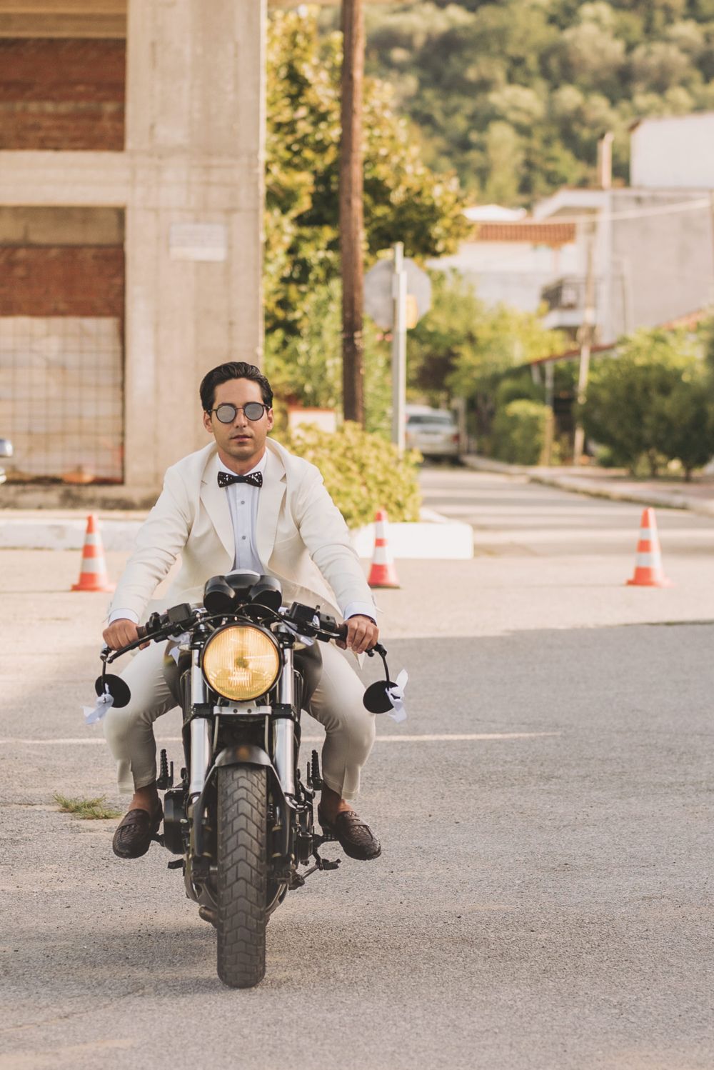 Stylishly dressed motorcycle rider in bow tie and glasses sits atop vintage cafe racer bike on urban street.