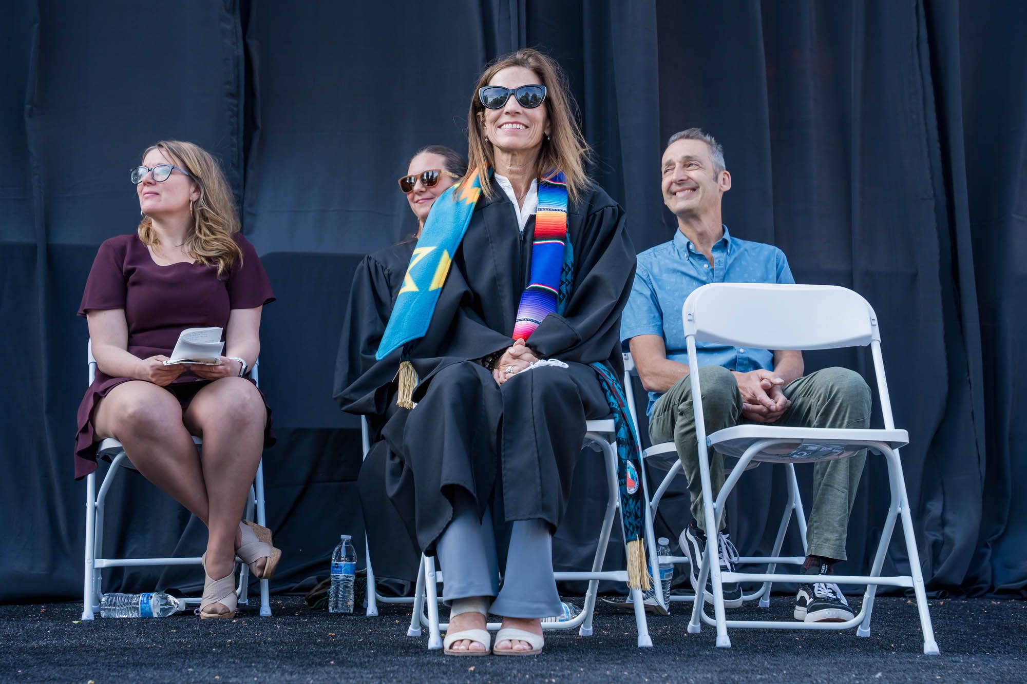 Four people sit on a row of white folding chairs against a black curtain background at an outdoor event.