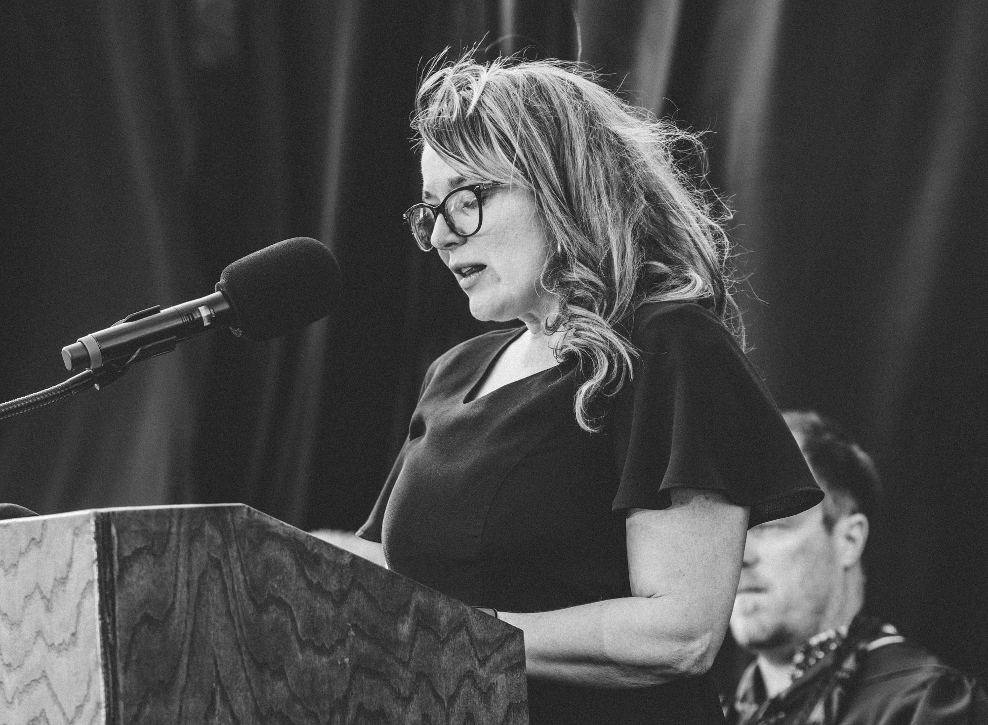 A black and white photo shows a performer speaking at a podium with a microphone while wearing glasses and a dark outfit.
