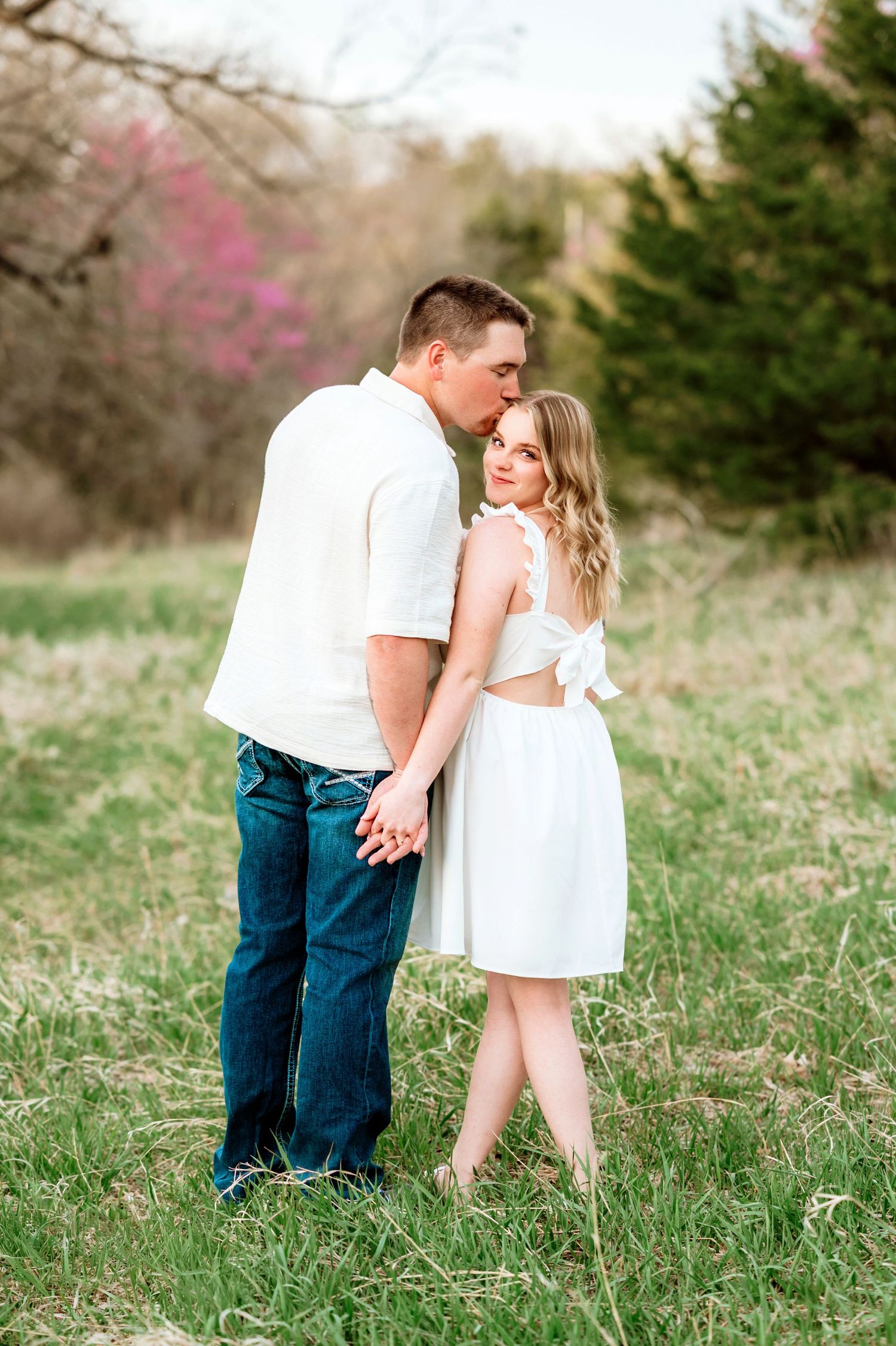 Romantic couple embraces in a spring meadow with blooming trees in the background wearing white outfits.