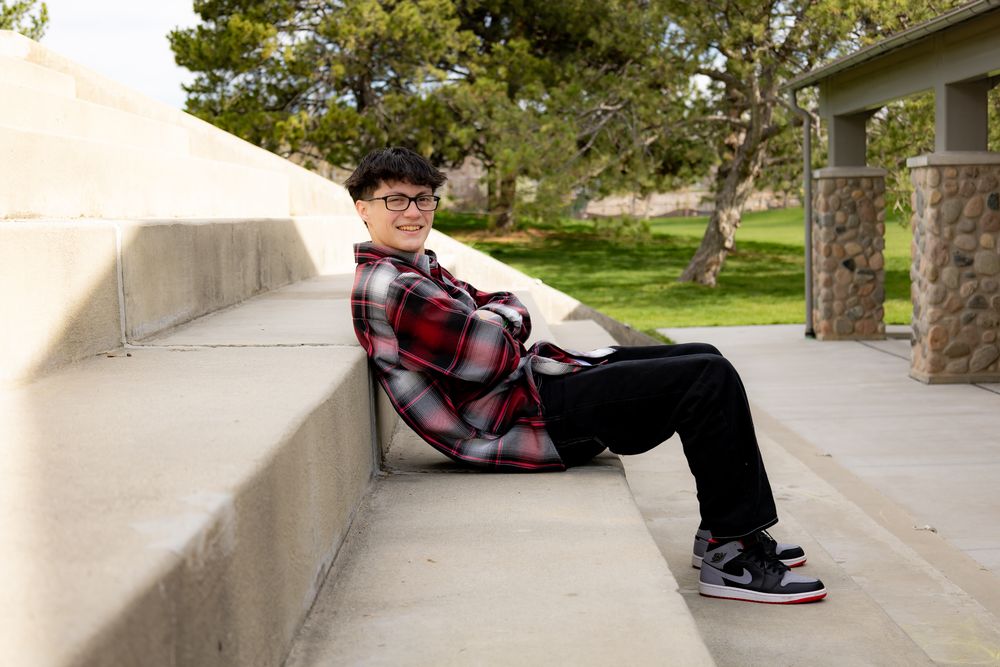 Graduate in plaid shirt and black pants relaxing on concrete steps outdoors.