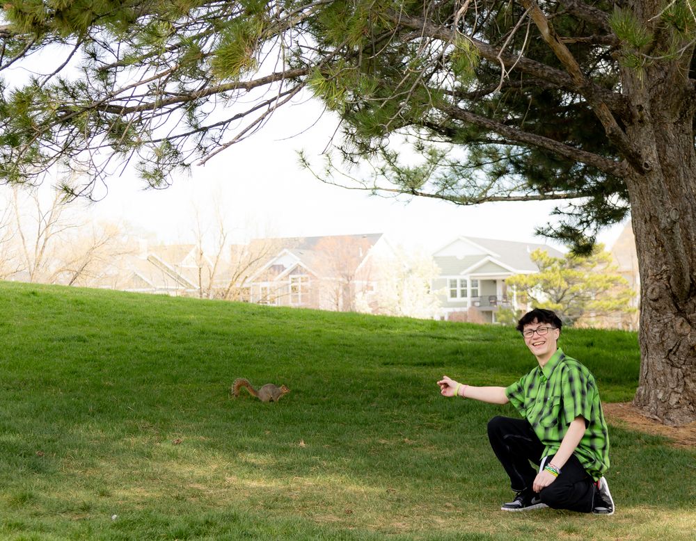 Graduate in green plaid shirt kneeling on grassy hill with houses in background.