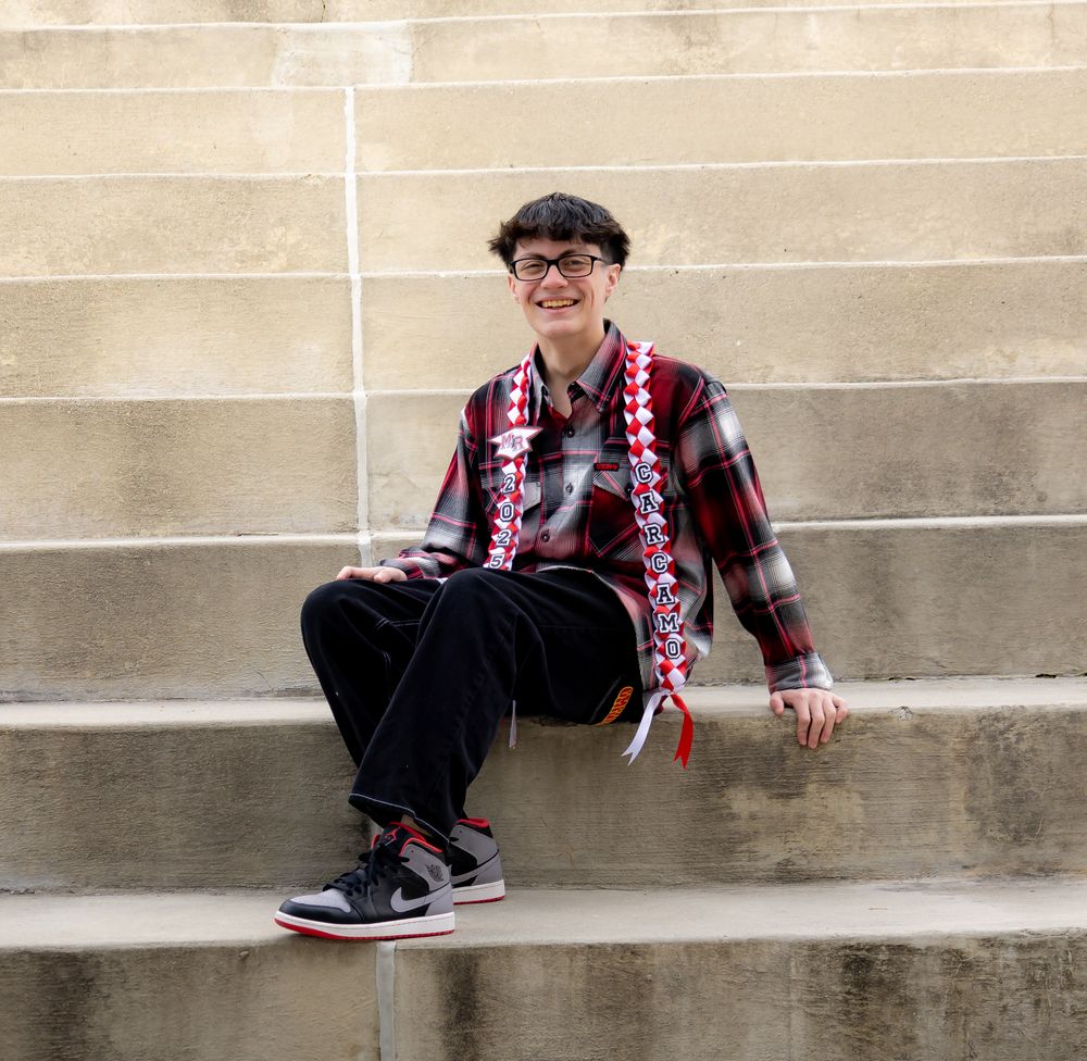 Graduate in festive plaid shirt with decorative sash sitting on concrete steps.