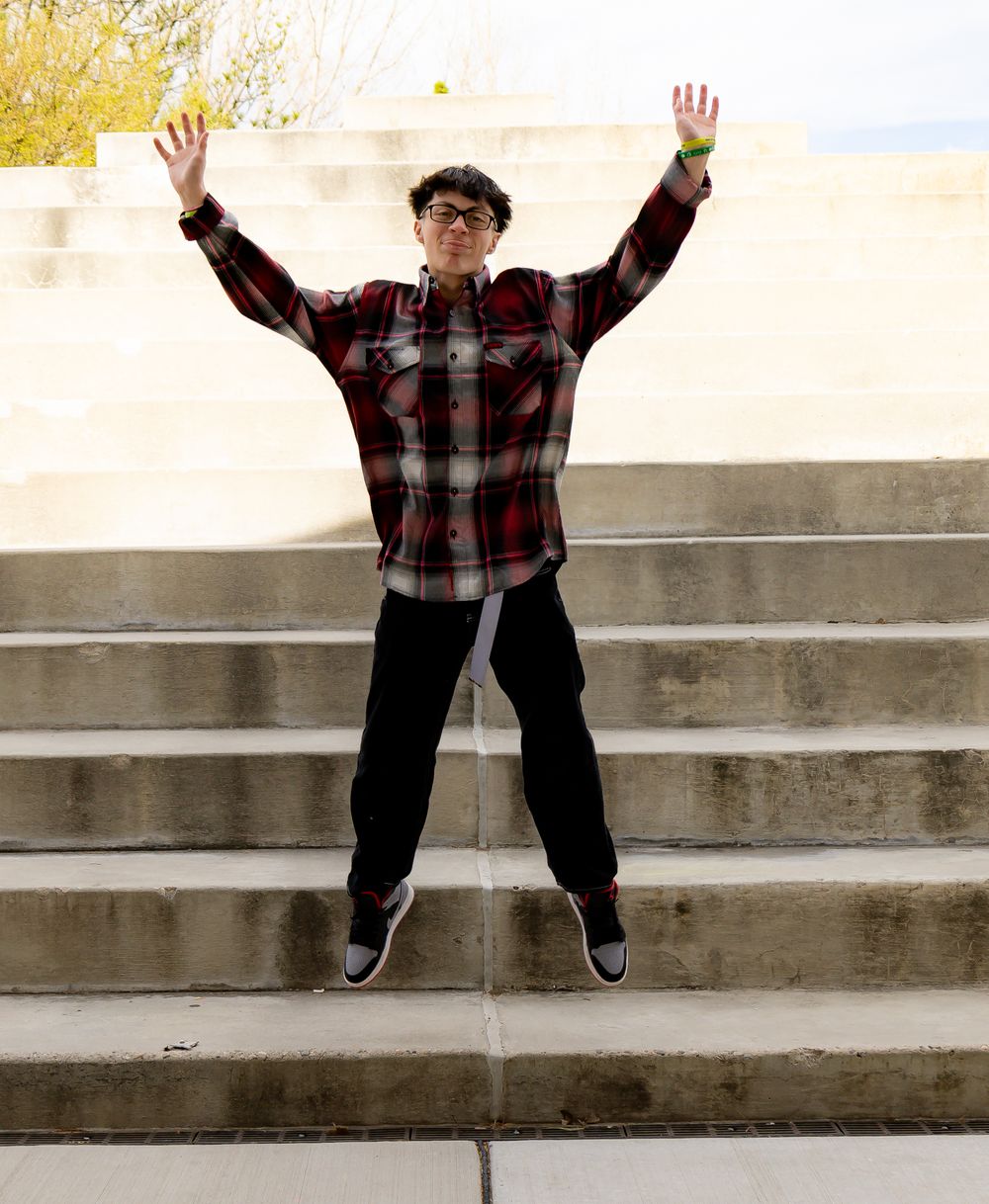 Graduate in plaid shirt and black pants jumping joyfully on outdoor steps.