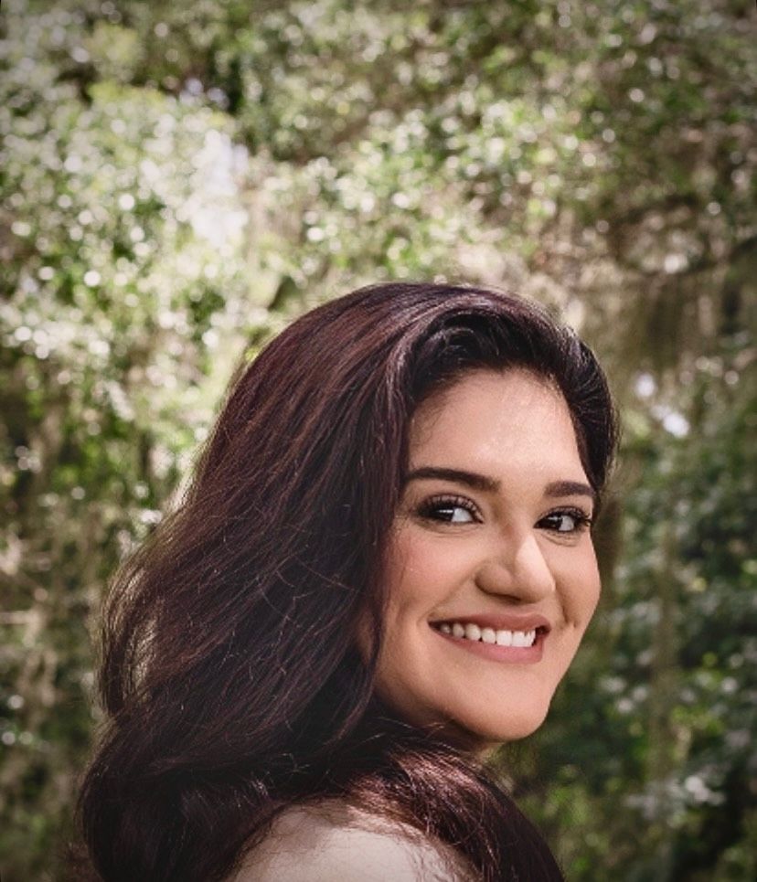 Young woman with dark wavy hair smiling beautifully in front of a lush green outdoor background.