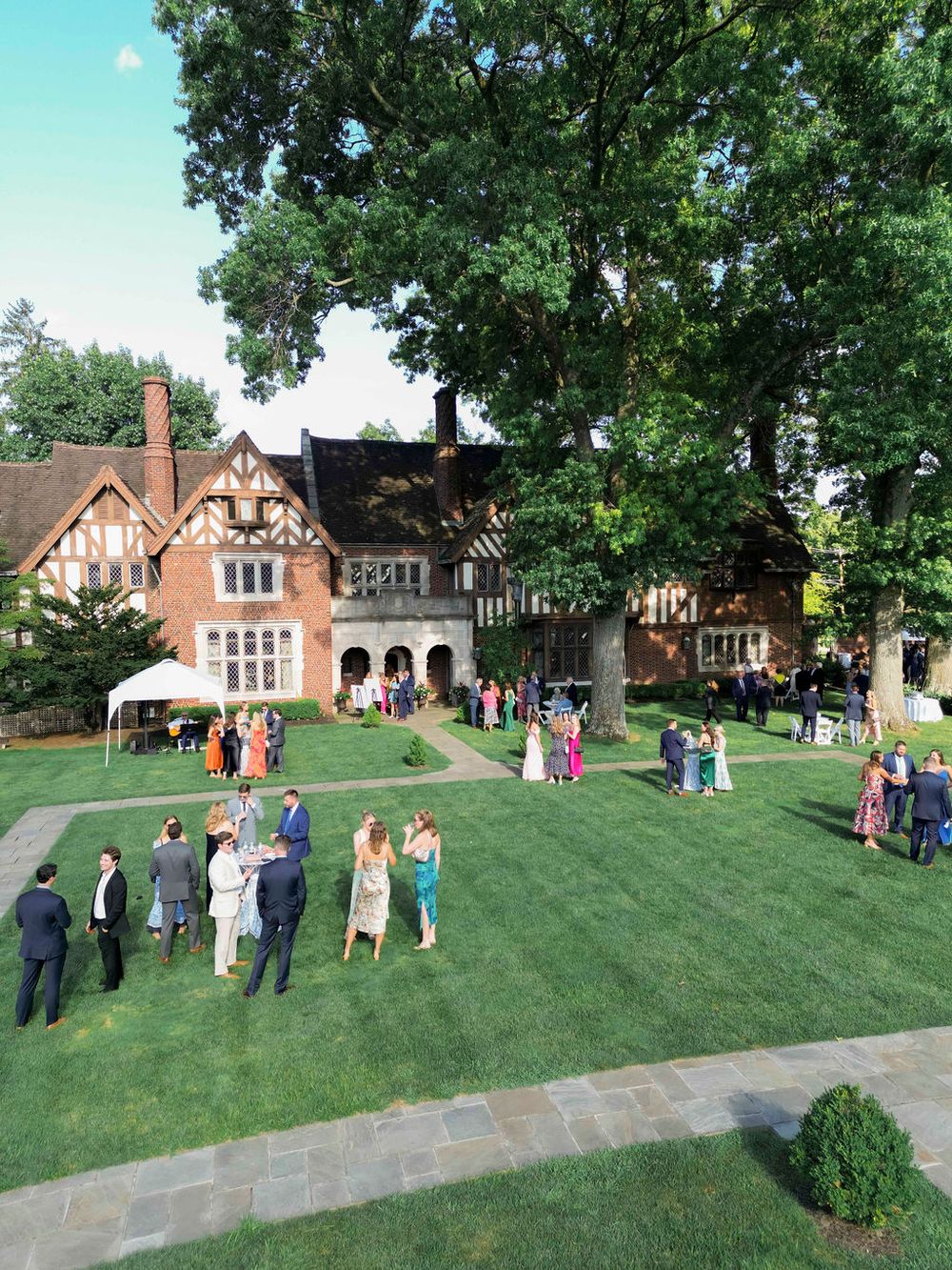 Wedding guests gather on a manicured lawn in front of an elegant Tudor-style brick mansion on a sunny day.