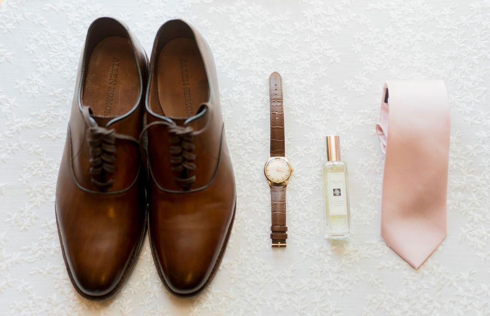 Wedding accessories including brown leather dress shoes, a wristwatch, cologne, and blush pink necktie laid out on white surface.