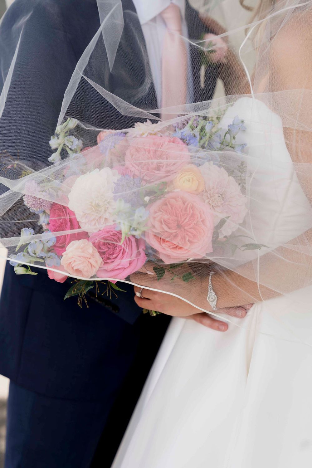 Beautiful wedding bouquet with pink garden roses and delicate tulle veil held between bride and groom in formal attire.