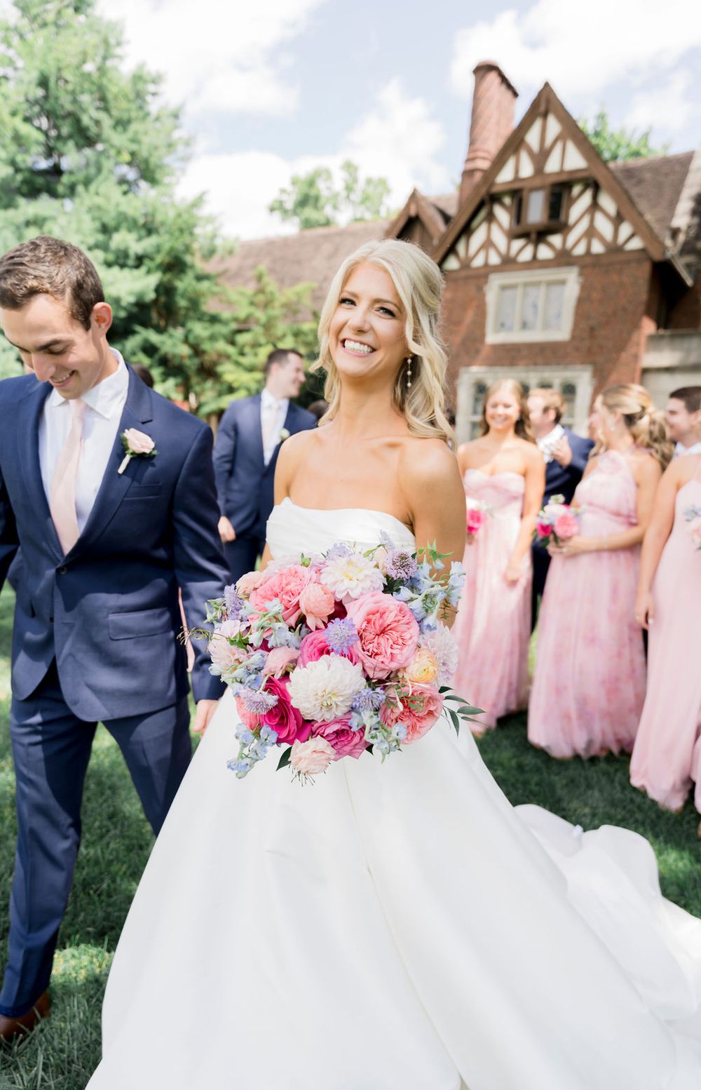 A wedding party poses on a lawn in front of a Tudor-style home with bridesmaids wearing pink dresses.