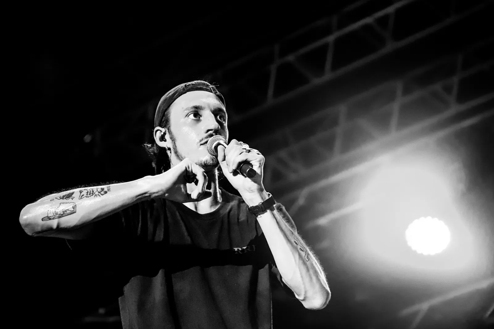 Rappeur en pleine performance live, bras levé, avec un projecteur intense derrière lui.