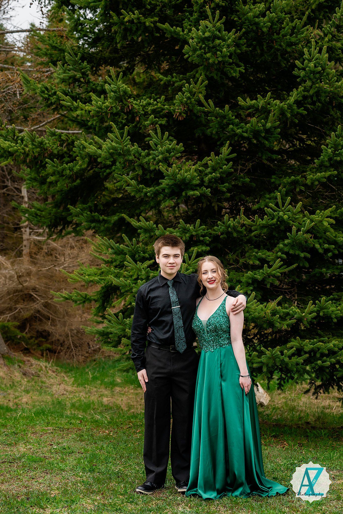 A couple in formal attire poses together in front of evergreen trees, with the woman wearing a flowing emerald green gown.