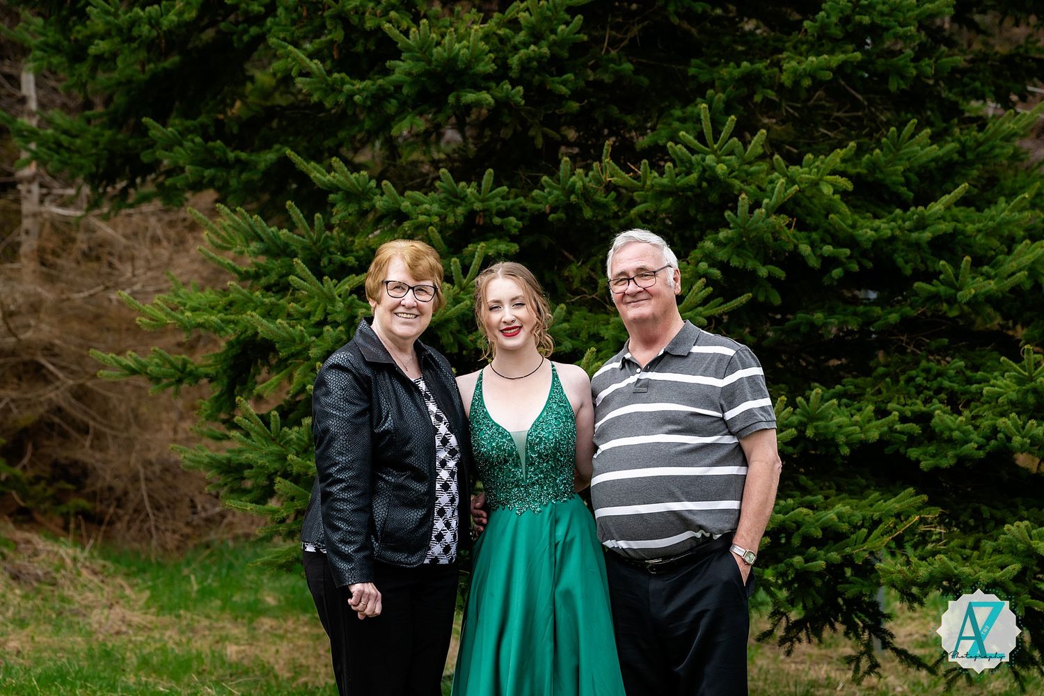 Three people pose together for a photo in front of evergreen trees.