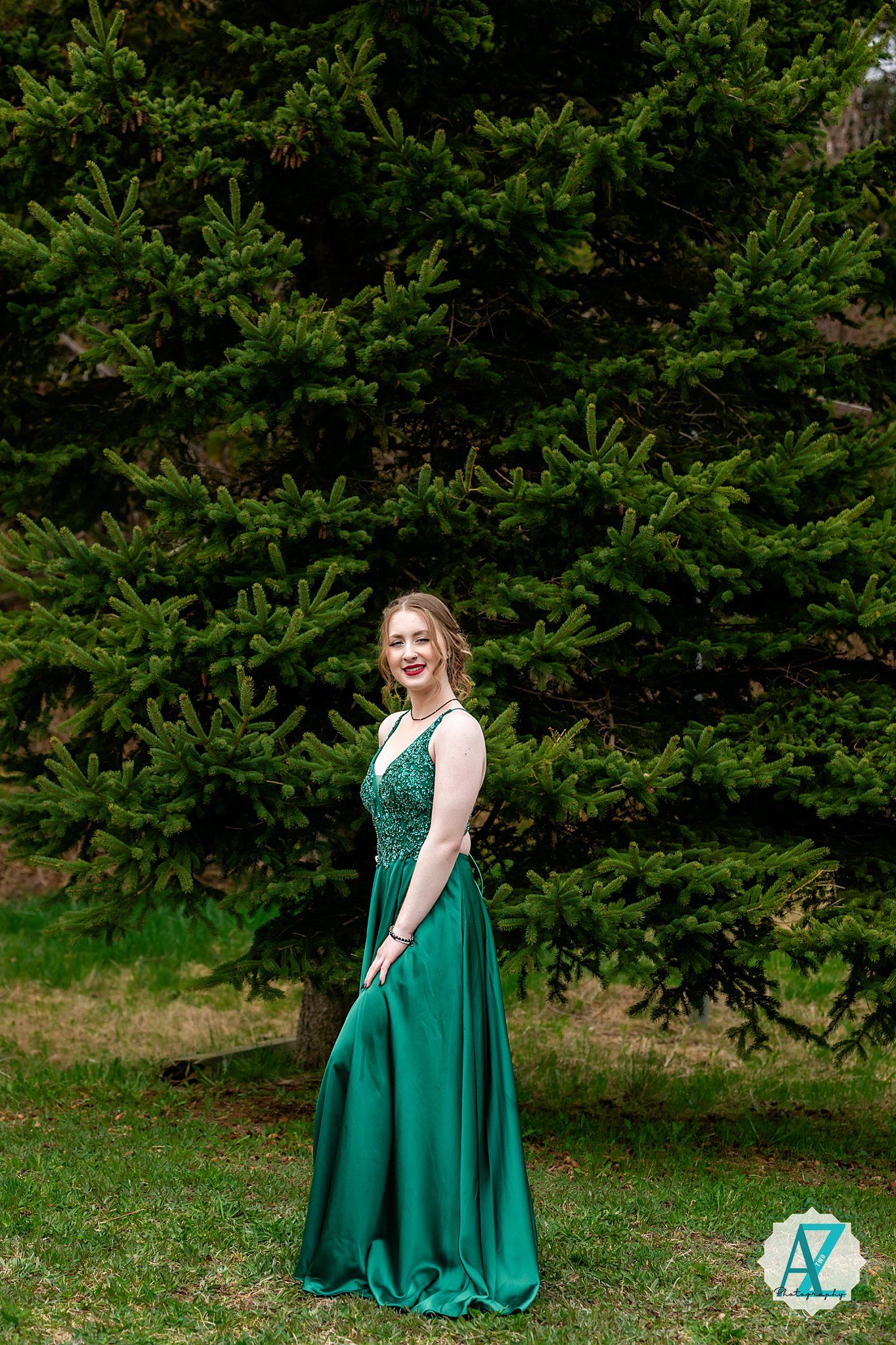 A person in an elegant emerald green formal gown poses in front of evergreen trees on a grassy lawn.