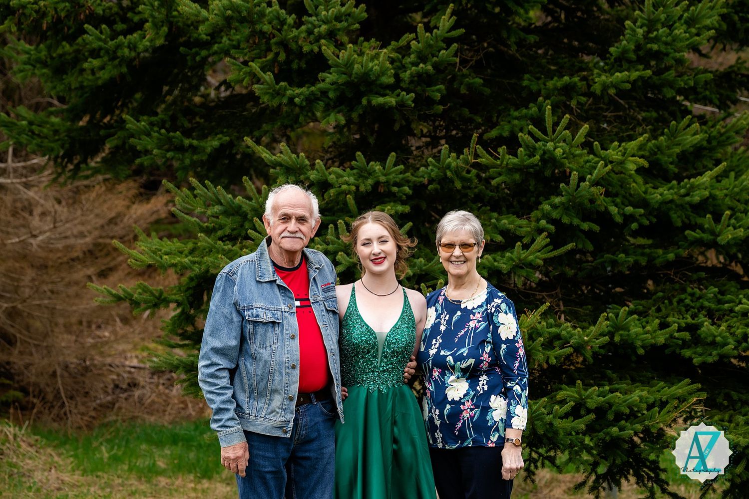 Three people stand together outdoors in front of evergreen trees, with a person in a green dress in the center.
