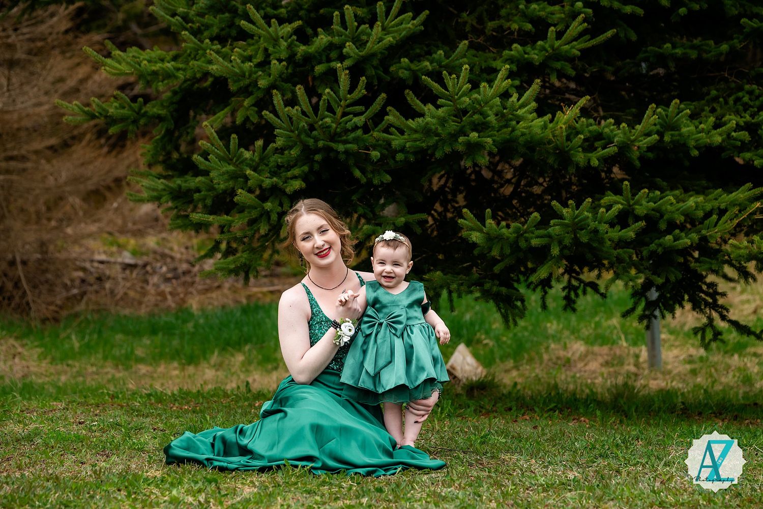 A pair in matching green dresses sit together on grass near evergreen trees in an outdoor portrait setting.
