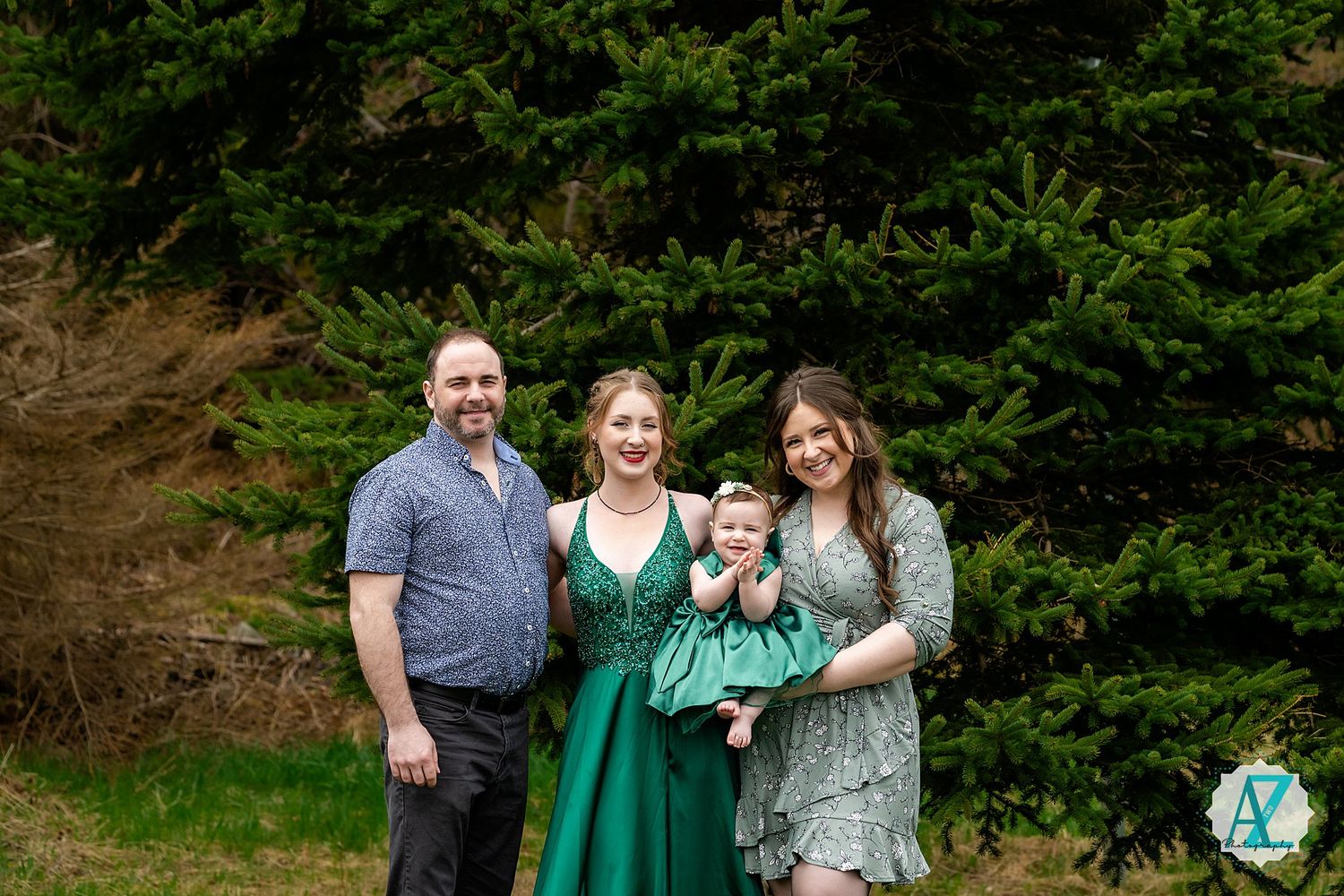 A family of four poses together outdoors in front of evergreen trees, with two wearing green clothing.