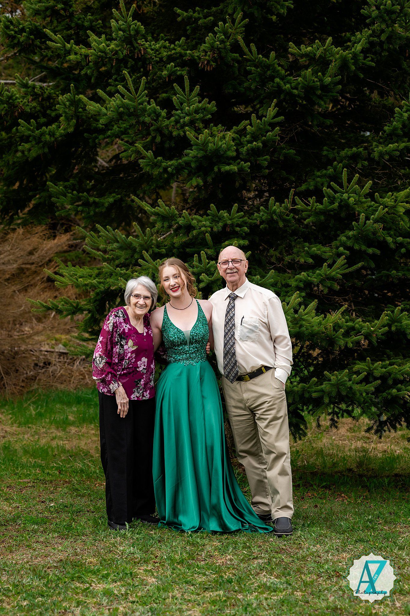 Group photo against evergreen trees with one person wearing an elegant emerald green gown at an outdoor event.