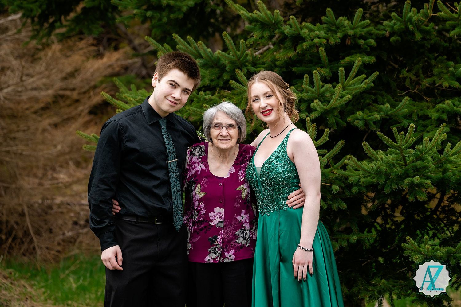 Three people pose together outdoors for a special occasion photo in front of evergreen trees.