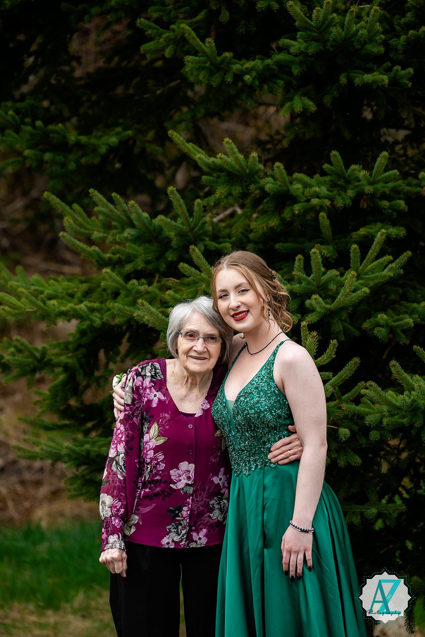 Two people stand together and smile in front of an evergreen tree, one wearing a burgundy floral top and one in emerald formal dress.