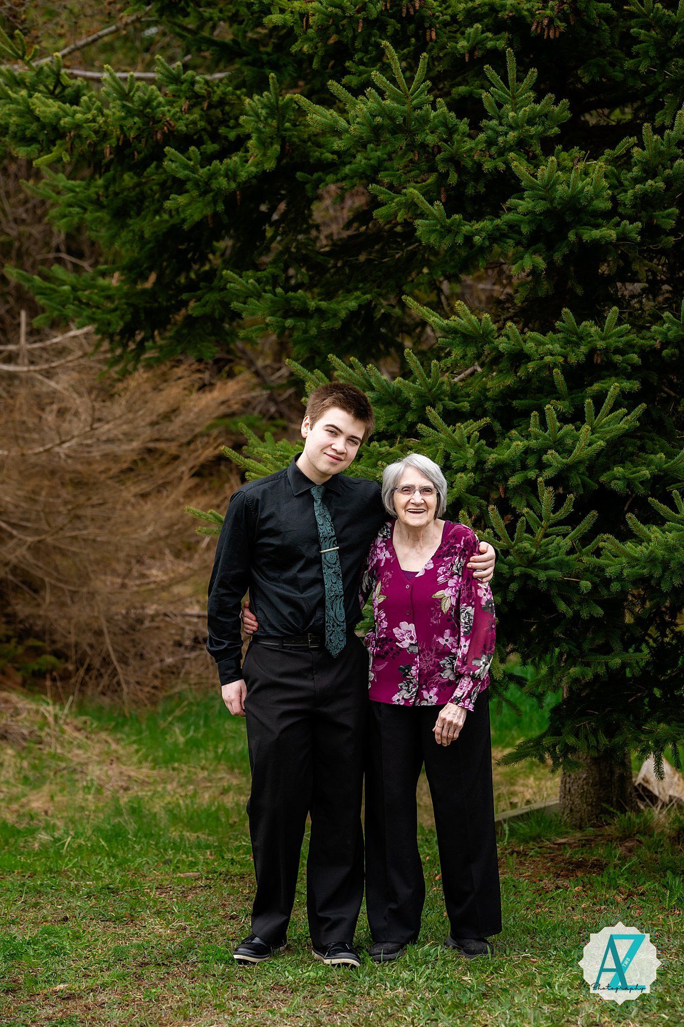 Two people pose for a formal portrait in front of pine trees on a grassy slope.