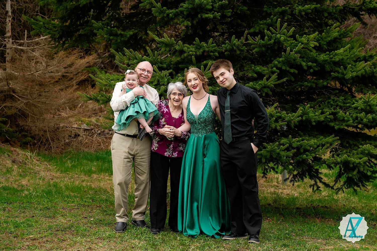 A group poses together outdoors on a grassy area with evergreen trees in the background.