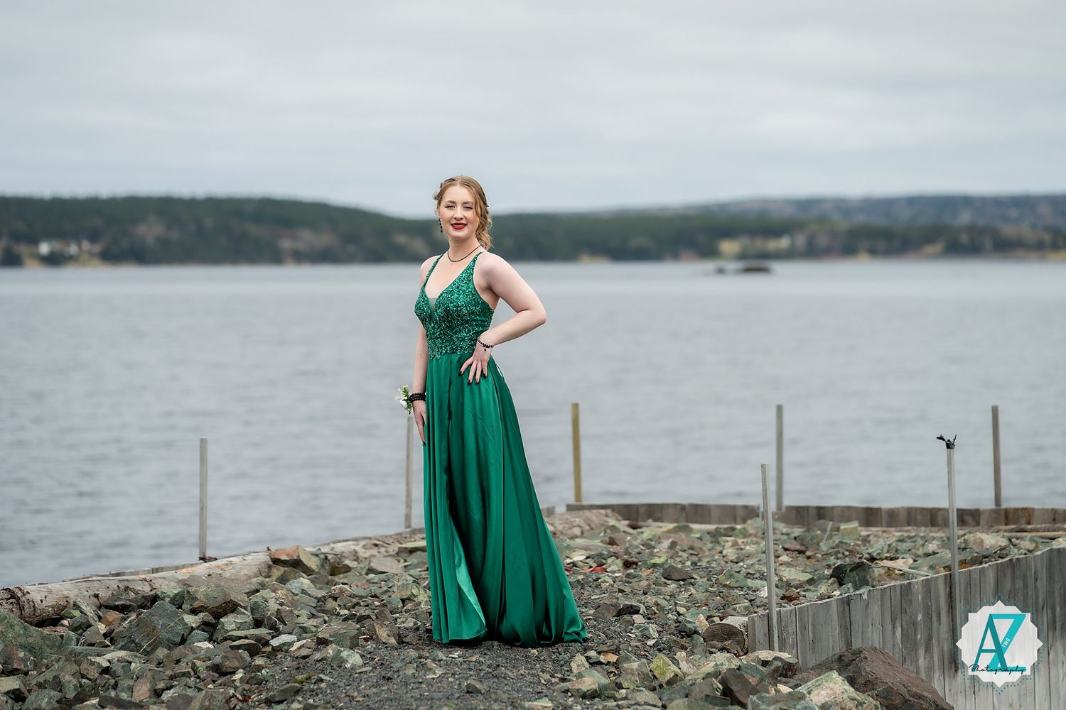 Figure in an elegant emerald green gown stands on rocky coastal shoreline with water and hills in background.