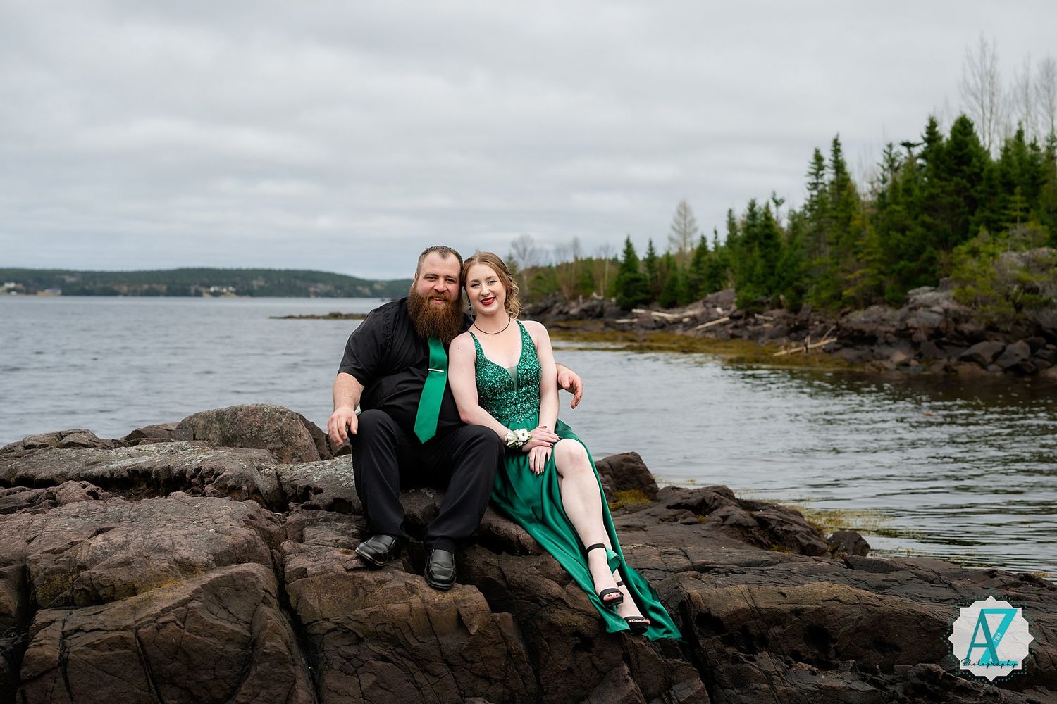 Couple sits together on rocky coastline overlooking scenic water and forested hills.