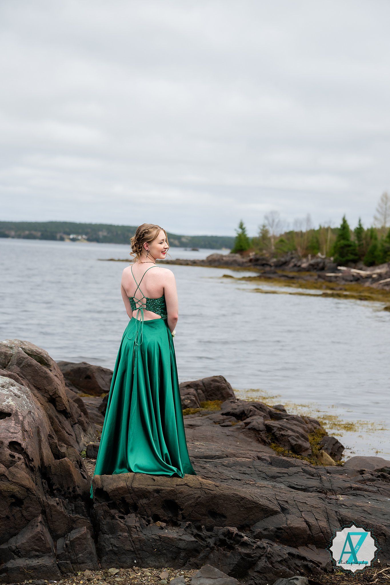 An emerald green formal dress flows in the wind as someone stands on rocky coastal cliffs overlooking a serene lake.