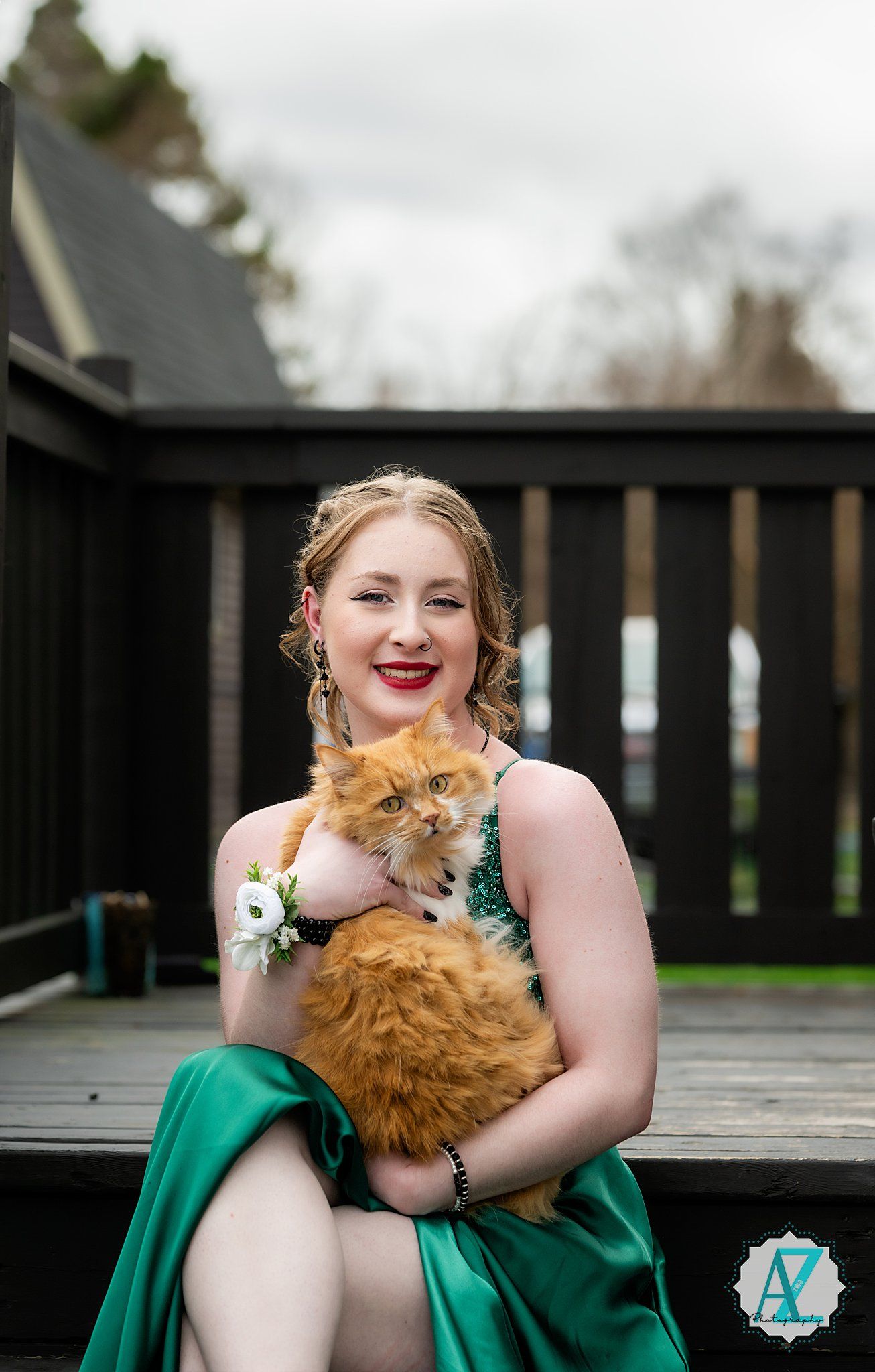 A person in an elegant emerald green formal dress holds a fluffy orange Persian cat while sitting on a wooden deck.