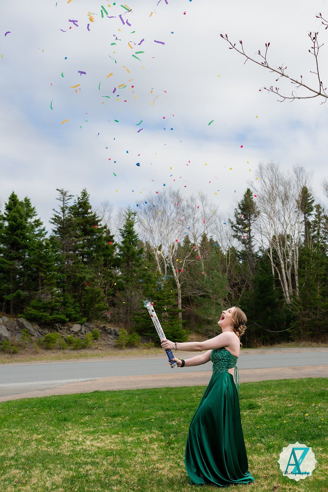 A person in an emerald green formal gown releases colorful confetti into the air against a backdrop of pine trees.